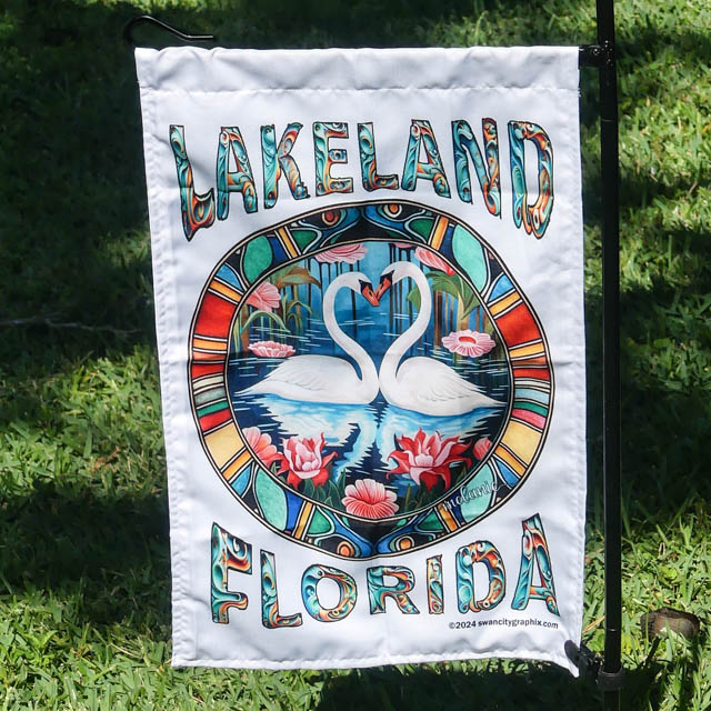 A hanging 12 x 18 Art Deco Lakeland Florida Swan City Garden Flag with an artistic illustration reading Lakeland Florida in a stained-glass style. It features two white swans forming a heart shape with their necks, swimming among pink water lilies and green foliage, with a sunset sky in the background. The text and frame have vibrant, colorful patterns in garden