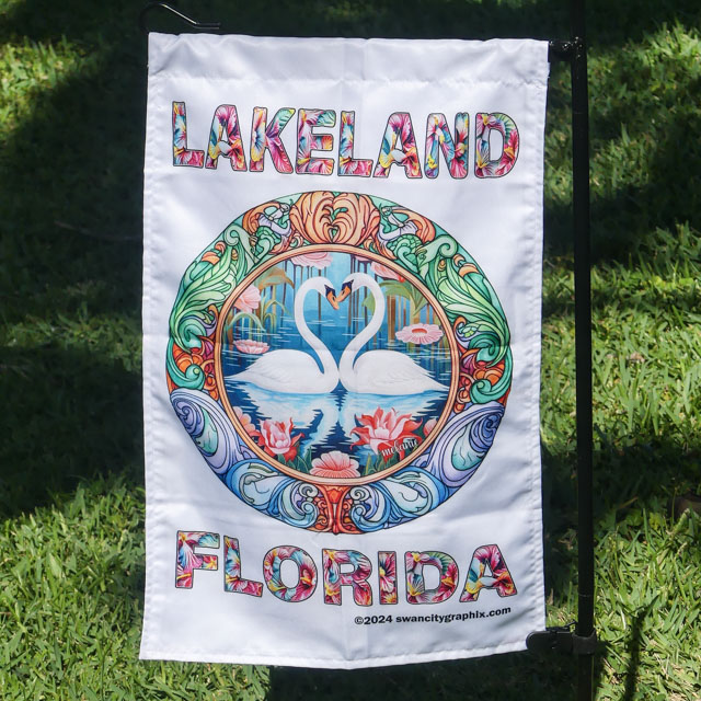 Vivid 12x18 Garden Flag hanging outdoors, illustrated text LAKELAND FLORIDA surrounds a circular image of two swans forming a heart shape with their necks over water, with pink water lilies and lily pads. Intricate, colorful patterns in an Art Nouveau design frame the scene | Swan City Graphix