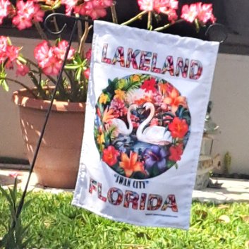 Floral illustration featuring a pair of white swans swimming in a vibrant, tropical setting surrounded by lush flowers and greenery. LAKELAND and FLORIDA are written in large letters decorated with floral patterns, and SWAN CITY is written beneath the illustration for this Swan City Garden Flag lifestyle pic hanging on wire stand in garden