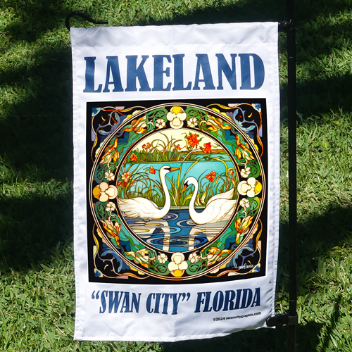 Art Nouveau 12x18 inch Garden Flag by Swan City Graphix with colorful illustration featuring two white swans on a blue pond surrounded by grass and orange flowers. The Art Nouveau border is intricately designed with floral patterns. The text LAKELAND is displayed at the top, and SWAN CITY FLORIDA is written at the bottom, lifestyle pic hanging on wire stand in garden